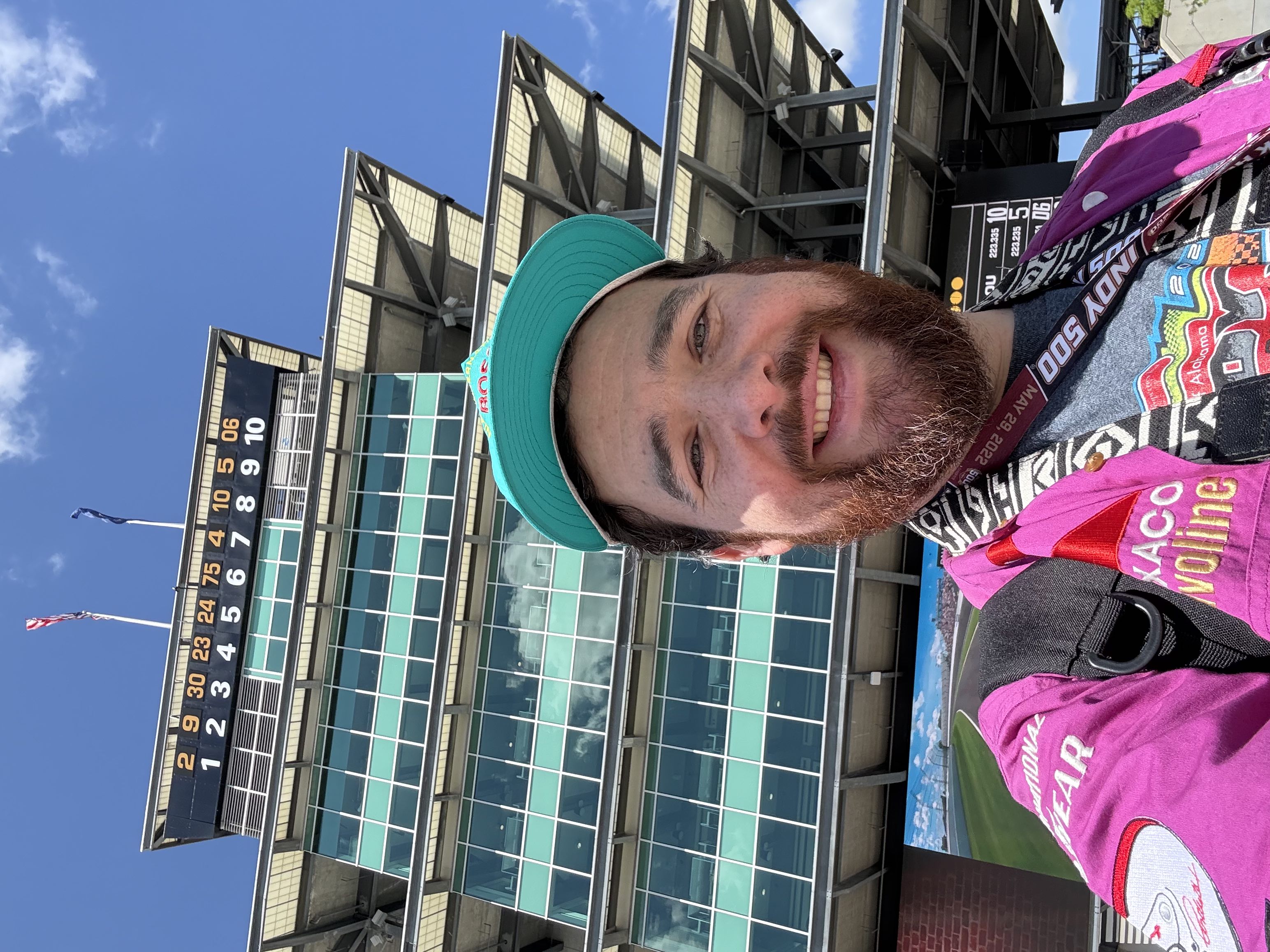 A man in colorful motorsports clothes smiling for a selfie with the pagoda control tower at Indianapolis Motor Speedway behind him
